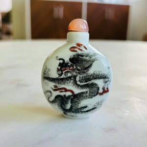 May include: A small, round, white ceramic snuff bottle featuring a hand-painted black dragon with red accents. The bottle has a smooth, glossy finish and is topped with a rounded, translucent, pale pink stopper that fits into the narrow neck.
