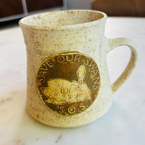 May include: A speckled beige ceramic mug with a curved handle. The mug features a circular emblem with a swan illustration and the text "SAVE OUR SWANS S.O.S."