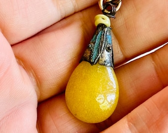 May include: A vintage pendant with a teardrop-shaped yellow stone. The pendant has a silver-colored metal cap with teal and black accents, and a gold-colored loop at the top for hanging. The stone is a vibrant yellow color.