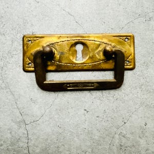 Op de afbeelding: Antieke messing ladeknop met een sleutelgatontwerp. De rechthoekige achterplaat heeft sierlijke details en een gebogen handgreep. De hardware heeft een warme, verouderde goudkleur, wat een vintage charme suggereert. Het sleutelgat is gecentreerd.