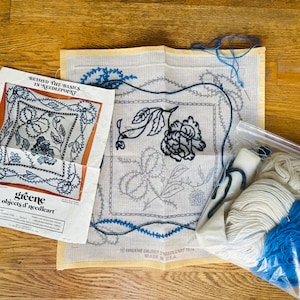 以下が含まれることがあります： クリーム色のキャンバスに青と黒の糸を使った花柄のデザインのニードルポイントキット。パターン、糸、部分的に完成したニードルポイント作品が含まれています。「greene objects d' needleart」と「MADE IN U.S.A.」と記載されています。