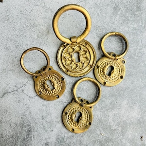 Może przedstawiać: Cztery antyczne mosiężne szyldy z otworami na klucz z dołączonymi uchwytami pierścieniowymi. Szyldy mają okrągły kształt z otworem na klucz pośrodku i dekoracyjnymi detalami na krawędziach. Uchwyty pierścieniowe są okrągłe i przymocowane do górnej części szyldów.