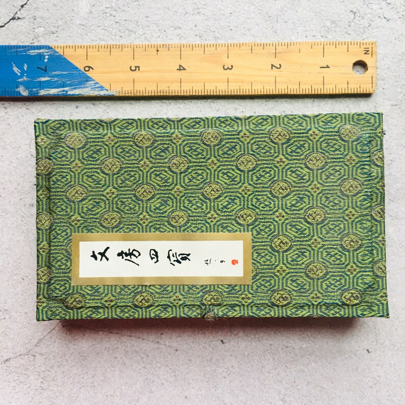 May include: A small, rectangular green box with a geometric pattern, a white label with Japanese text, and a red stamp. A blue and yellow ruler is placed above the box, showing measurements in inches.