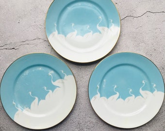 May include: Three ceramic plates with a light blue and white design. The plates feature a gold rim and a pattern of white birds against a light blue background. The plates are arranged on a gray surface.