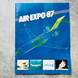 Può includere: Brochure blu con la scritta "AIR EXPO 87" in alto. La brochure presenta un'illustrazione a filo di un aereo e quattro immagini più piccole in basso. Le immagini includono un aereo, una tuta spaziale e altri velivoli.