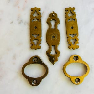 Puede incluir: Una colección de herrajes antiguos de latón, que incluyen tiradores y escudos decorativos. Los herrajes presentan diseños ornamentados y un acabado cálido y envejecido. Las piezas están dispuestas sobre una superficie de mármol.