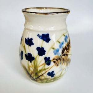 Puede incluir: Un jarrón de cerámica de color crema con un borde marrón oscuro. El jarrón está decorado con diseños florales azules y celeste con tallos verdes. El jarrón tiene una forma redondeada, ideal para exhibir flores. Altura aproximada 15 cm.