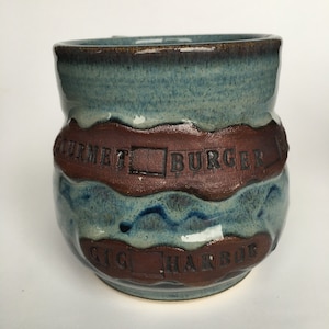Puede incluir: Una taza de cerámica azul y marrón con las palabras "Gourmet Burger" y "Gig Harbor" estampadas en ella.