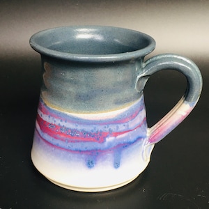 May include: A handmade ceramic mug with a unique design. The mug features a dark blue rim, transitioning to a band of purple and red, and a white base. The handle is a continuation of the mug's design. This mug is perfect for coffee or tea.