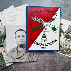 May include: A vintage book cover with the text "Black Hawks Air Cavalry Squadron 7/1st 1971" and an illustration of a hawk and helicopters. The cover is red, white, and blue. There are also black and white photos and documents.
