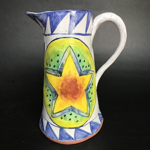 Puede incluir: Una jarra de cerámica blanca con un diseño de estrella azul y verde. La jarra tiene un asa y un pico. La estrella es amarilla con un centro rojo.