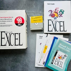 Op de afbeelding: Een Microsoft Excel versie 5.0 softwarepakket voor Windows, inclusief een grote witte doos met rode prijstekst, een 3,5-inch diskette met een gele registratiesticker, een witte gebruikershandleiding en een blauwe Microsoft Excel resourcegids.