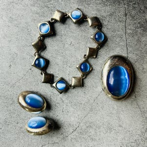 May include: A silver bracelet with blue and white stones, a matching brooch, and a pair of earrings. The jewelry is made of silver and has a vintage look.