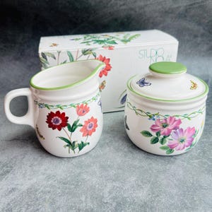 May include: A white ceramic creamer and sugar bowl set with a floral and butterfly design. The creamer has a handle and a green rim. The sugar bowl has a lid with a green knob. The set is in front of a box with a similar design.