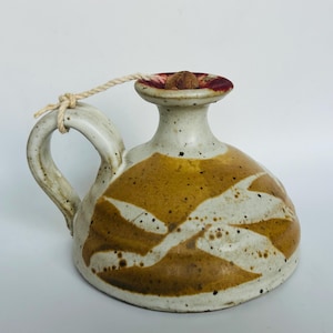 Puede incluir: Jarra de cerámica hecha a mano con asa y boquilla. La jarra presenta una base color crema con patrones abstractos marrones y blancos. El borde es rojo y una cuerda natural está unida a la tapa. La jarra mide aproximadamente 15 cm de altura.