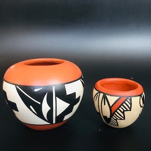 Könnte beinhalten: Zwei handbemalte Keramiktöpfe. Der größere Topf ist orange mit schwarz-weißen geometrischen Mustern. Der kleinere Topf ist beige mit orangefarbenen, schwarzen und weißen Mustern. Beide Töpfe haben eine glatte Oberfläche und stehen vor schwarzem Hintergrund.