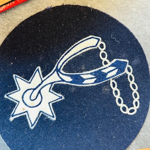 May include: A circular dark navy blue felt patch featuring an embroidered white and navy blue spur design. The spur includes a star-shaped rowel and a chain detail, all rendered in clean, contrasting thread work against the solid dark background.