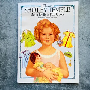 May include: Classic Shirley Temple Paper Dolls in Full Color book cover featuring a color illustration of Shirley Temple holding a doll. The book cover has a blue border and the title is in white text.
