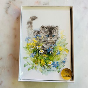 以下が含まれることがあります： 青い目をした灰色の猫の絵が描かれたグリーティングカードの箱入りセット。青、黄、白の花の中にいます。子猫が焦点で、緑に囲まれています。リサイクル紙に印刷されています。