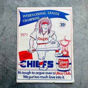 May include: Vintage Syracuse Chiefs baseball poster from 1971, featuring a cartoon Native American baseball player. The poster includes text that reads "International League Champions" and "Junior World Series Champs". The poster also has the text "It's tough to argue over a Utica Club."