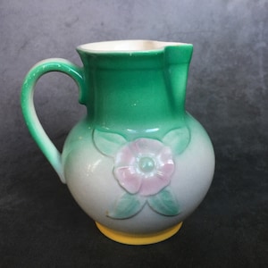 Può includere: Un'anfora in ceramica verde e bianca con un motivo floreale rosa. L'anfora ha una base gialla e un manico.