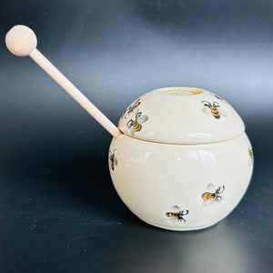 May include: Cream-colored ceramic honey pot with a lid and a wooden honey dipper. The pot is decorated with black and yellow bee illustrations. The dipper is inserted into the pot. The honey pot is round and has a lid.