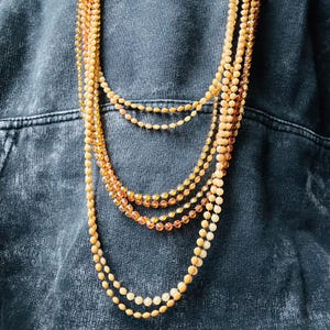 May include: A layered necklace with multiple strands of small, round beads. The beads are in shades of beige and amber. The necklace is displayed against a dark denim background. The beads are strung together, creating a layered effect.