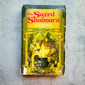 Puede incluir: Libro de bolsillo vintage titulado "The Sword of Shannara" de Terry Brooks. La portada presenta una pancarta amarilla con el título en rojo y una ilustración de fantasía de tres figuras con una espada brillante. El libro tiene un aspecto desgastado.