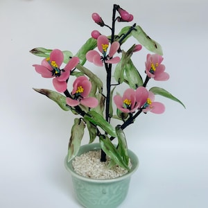 May include: A glass flower arrangement in a green ceramic pot. The flowers are pink with yellow centres and green leaves. The pot is filled with white gravel.