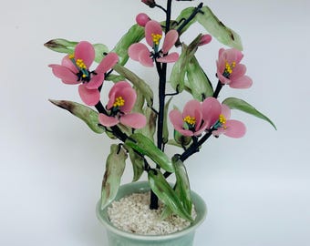 May include: A glass flower arrangement in a green ceramic pot. The flowers are pink with yellow centers and green leaves. The pot is filled with white gravel.