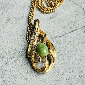 May include: A gold-toned necklace featuring a pendant with an oval, light green stone. The pendant has an ornate, swirling design. The chain is also gold-toned and visible at the top of the image. The necklace is displayed on a gray surface.