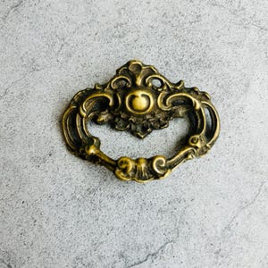 May include: Ornate antique brass drawer pull with a decorative design. The pull features a circular handle and intricate detailing, perfect for adding a vintage touch to furniture. The brass finish has a warm, aged appearance.