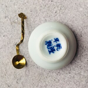May include: A small, white ceramic teacup with blue Chinese characters on the bottom, paired with a decorative gold-colored spoon. The spoon has a cloud-shaped handle and a round bowl. The teacup and spoon are set against a neutral background.