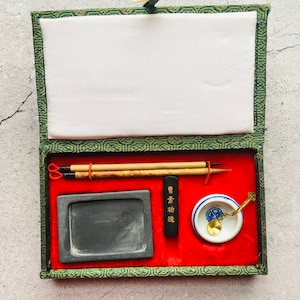 May include: A calligraphy set in a green and red box. The set includes two bamboo brushes, a rectangular ink stone, a black ink stick with gold text, a small ceramic dish, and a gold-colored spoon. The box is open, revealing a white fabric lining.