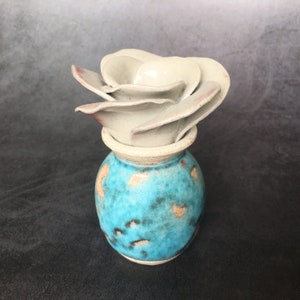 May include: A small, turquoise ceramic vase with a white, ceramic flower on top. The vase has a speckled, blue and brown glaze.