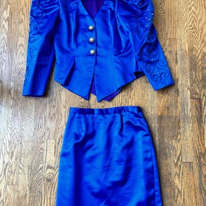 May include: A vibrant blue satin skirt suit. The jacket features decorative embroidery on the sleeves, shoulder pads, and gold buttons. The skirt is a matching knee-length pencil skirt. The suit is displayed on a wooden surface.
