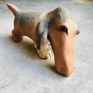 May include: A ceramic dachshund ornament in brown, grey, and cream. The dog has a long snout, floppy ears, and a slightly upturned tail. The figurine is handmade and has a rustic, artistic appearance.
