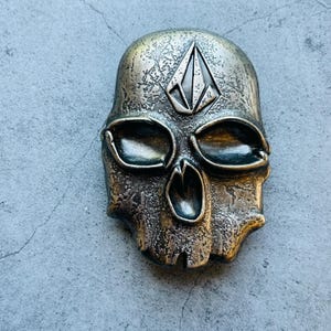 May include: A metallic skull-shaped belt buckle with a textured surface and a diamond-shaped logo on the forehead. The buckle has a bronze and silver color scheme, with detailed eye sockets and a mouth opening.