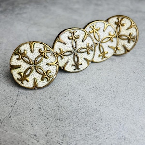 May include: A set of four circular buttons featuring a textured white enamel surface with an embossed antique gold-toned metal fleur-de-lis cross pattern. Each button has a raised metallic rim and a repeating decorative design across the face.