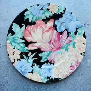 May include: A round black plate featuring a floral pattern. The flowers are in shades of pink, blue, and cream, with green foliage. The plate has a gold rim and is set against a grey background.