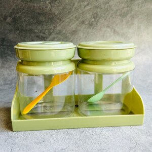 Puede incluir: Un juego de dos botes de especias transparentes con tapas y bases de color verde claro, cada uno con una cuchara pequeña. Los botes están en una bandeja verde claro. Una cuchara es amarilla, la otra verde.