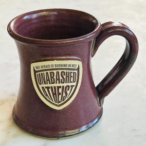 Puede incluir: Una taza de cerámica burdeos con un asa curvada. La taza presenta un escudo color crema con las palabras "NOT AFRAID OF BURNING IN HELL" encima de "UNABASHED ATHEIST" en una fuente audaz y estilizada. La taza tiene un acabado brillante.