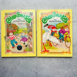 Könnte beinhalten: Zwei Vintage-Geschichtenbücher von Cabbage Patch Kids mit gelben Umschlägen. Das Buch links trägt den Titel "The Just-Right Family" und das Buch rechts den Titel "The Big Bicycle Race". Beide Bücher sind mit Figuren aus der Cabbage Patch Kids-Reihe illustriert.