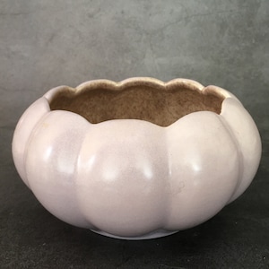 Könnte beinhalten: Eine hellrosa Keramikschale mit gewelltem Rand. Die Schale hat eine kürbisartige Form mit vertikalen Rippen. Der Innenraum ist hellbraun. Die Schale steht auf einer dunklen Oberfläche.