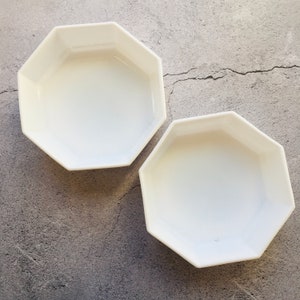 May include: Two white, octagonal ceramic bowls are displayed on a gray surface. The bowls have a simple, modern design, with a smooth, glossy finish. The bowls are empty and ready for use.
