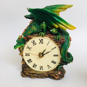 Puede incluir: Un reloj decorativo con un dragón verde posado sobre una base de madera falsa. La esfera del reloj es de color crema con números romanos negros y manecillas negras. El dragón tiene escamas verdes, alas y un acento rojo.