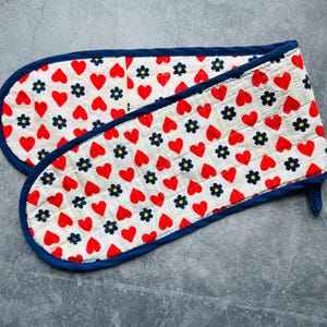 May include: A double oven mitt with a white background, red hearts, and blue flowers. The mitt has a blue trim and a loop for hanging. The pattern is repeated across the surface of the mitt.
