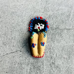 Könnte beinhalten: Handgefertigte puppenförmige Anstecknadel mit einem schwarzen, mit Perlen besetzten Kopf, roten und blauen Perlenbesatz und einem Körper aus hellbraunem Leder. Die Puppe hat ein weißes, mit Perlen besetztes Gesicht mit grüner Nase und blaue, rote und grüne Perlenakzente.