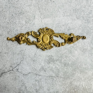 May include: Ornate, antique brass furniture hardware. The piece features a central oval design with a sunburst pattern and decorative scrollwork. Small, detailed faces are on each end. The hardware measures approximately 8 inches in length.