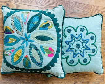 May include: Two square decorative pillows. One pillow features a central floral design with teal, blue, and multi-colored leaf-shaped appliqués. The other pillow has a starburst design. Both pillows have a teal background and dark green trim.